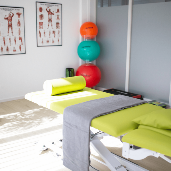 Ein heller Physiotherapieraum mit einem gelben Behandlungstisch, bunten Gymnastikbällen und anatomischen Postern an der Wand.
