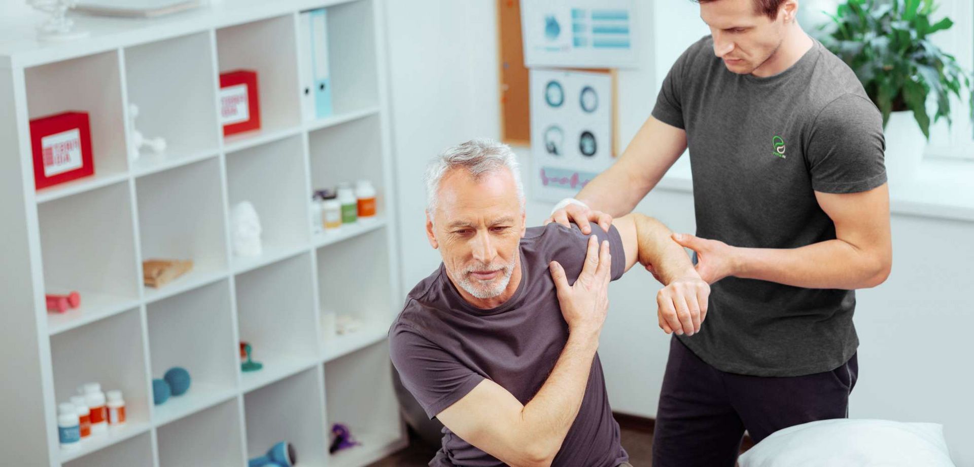 Ein Physiotherapeut hilft einem älteren Mann bei einer Armübung in einem hellen Behandlungsraum.