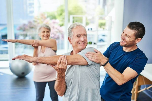 Ein Trainer unterstützt einen älteren Mann beim Dehnen, während eine Frau im Hintergrund ebenfalls Übungen macht.